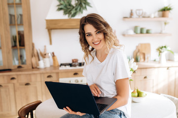 A smiling young girl with a laptop in her hands sits on a table in the kitchen. Freelancer