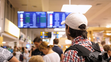 Young man with backpack checking flight information on digital schedule display inside arrival hall in international airport