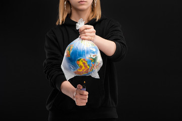 Partial view of woman holding lighter with fire under plastic bag with globe isolated on black, global warming concept