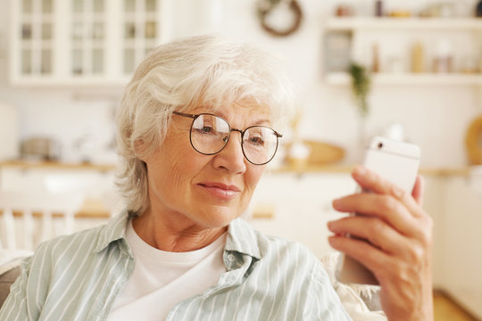 Attractive modern senior female pensioner in round glasses sitting on sofa, holding generic cell phone, reading sms. Retired gray haired woman browsing internet using 4 g wireless connection