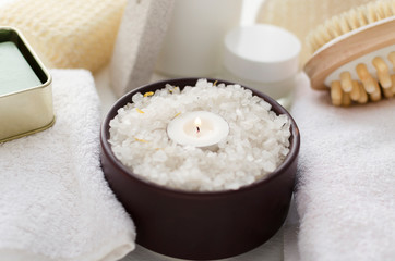 Brown bowl full of sea salt for bath procedures, candle flame, towels and other accessories for relaxation