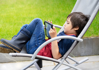 Obraz premium Portrait kid with playing game on tablet, A boy having fun watching cartoons on digital taplet, Child siting on roacking chair relaxing on weekend in the garden on spring or Summer