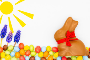 Happy easter card. Easter milk chocolate bunny, rabbit with red bow on a neck. There are colourful candies, flowers and sun. Copy space. 