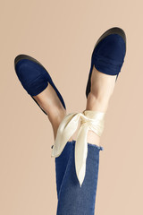 Close-up shot of crossed female legs in blue loafers and jeans on the beige background. One of the...