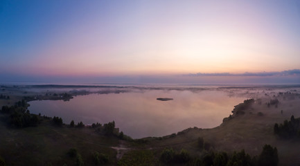 Misty sunrise over forest lake. Aerial panoramic view with trees, fog and golden sky.