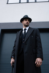 Fototapeta premium Low angle view of gangster in black coat and hat with weapon on street