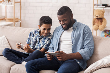 Cheerful Black Boy And His Dad Playing Video Games On Smartphones