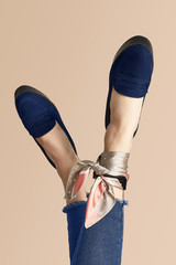 Close-up shot of crossed female legs in blue loafers and jeans on the beige background. One of the anklesis wrapped with a beige silk handkerchief with white dots and black and pink stripes. 