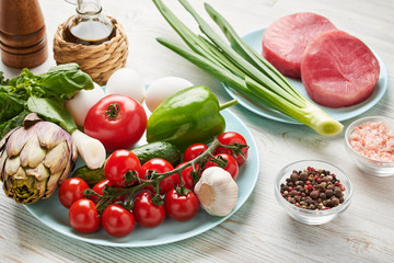 The ingredients for a classic tuna salad (Nicoise) lie on a blue plate. Artichoke, tomatoes, peppers, onions, fresh tuna, cucumber, garlic, basil, olive oil, eggs, salt, pepper.