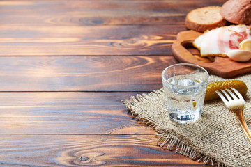 A glass of vodka and pickled cucumber on a fork. Fat with garlic and brown bread, on a wooden table. Close-up, place for text.