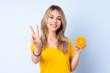 Teenager Russian girl holding an orange isolated on blue background smiling and showing victory sign