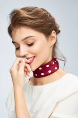 A chestnut-haired lady with careless hairdo in a white blouse is smiling and keeping her finger near the lips. A burgundy silk handkerchief with white polka-dot print is rounding the girl's neck.