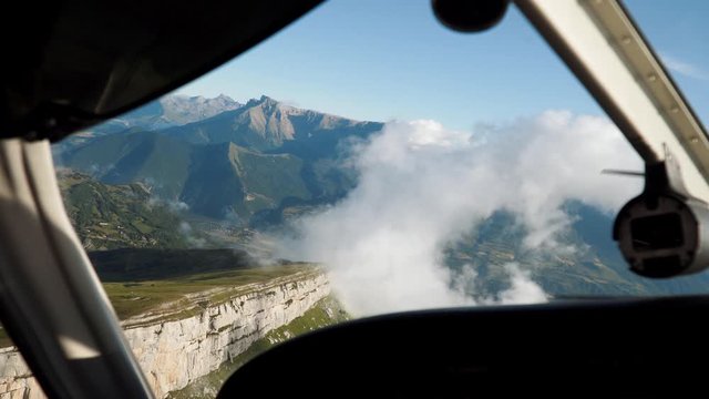 Flying small aircraft, cabin view. Clouds, mountains, cliff in airplane window. Driving private aeroplane high above rocks, moving through clouds in summer