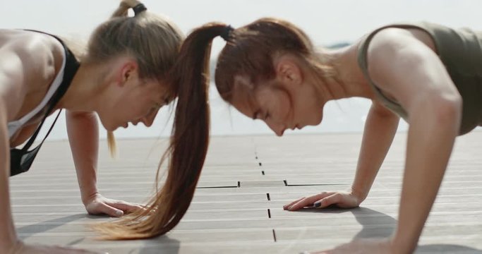 Two young woman doing push-ups on a wooden deck overlooking water head to head touching hands between each exercise in a health and fitness concept