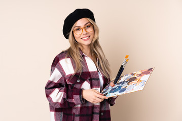 Teenager artist girl holding a palette isolated on blue background with glasses and smiling