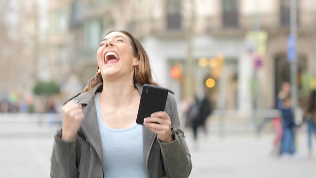 Excited young woman celebrating success checks phone in the street