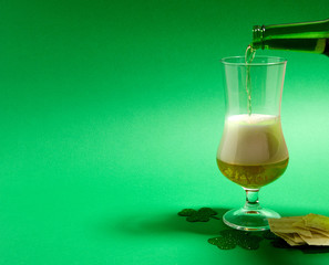 A glass of beer stands on a green background and a jet of beer from a green bottle. Nearby are chips and clover leaves.