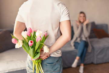 Young man hiding bouquet of tulips for girlfriend behind his back at home, blank space