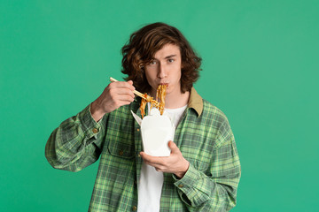 Food delivery concept. Young man eating takeaway noodles on color background, blank space for design