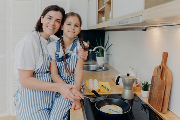 Lovely mother and daughter in aprons embrace and smile happily, fry eggs on modern stove in kitchen, use frying pan, prepare tasty breakfast. Family, children, motherhood and cooking concept