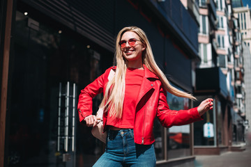 Well-dressed lady in good mood playfully posing on city background.Outdoor portrait of cute blonde lady wears elegant red jacket.