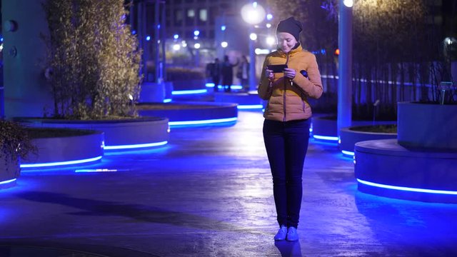 European woman watch to phone, then laugh, stand at night alley of linear park in Seoul. Blue illumination at walkway, modern Asian metropolis urban environment