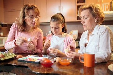 Multi-generational female Caucasian family preparing and decorating Easter eggs standing behind kitchen counter at home