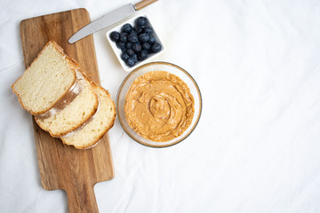 Top view of peanut butter in a bowl serving with bread and blueberries on white linen tablecloth for breakfast or picnic. Food concept for international peanut butter and jelly day with copy space