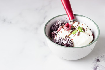 Vanilla ice cream with frozen raspberries, blackberries, chocolate and mint in a ceramic bowl, light grey background.