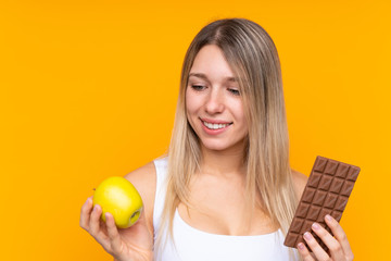 Young blonde woman over isolated blue background taking a chocolate tablet in one hand and an apple...