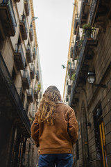 Naklejka premium Blond girl walking through the streets of the center of the city of Barcelona in Catalonia, Spain wearing a jacket and blue jeans. Looking up to the old buldings.