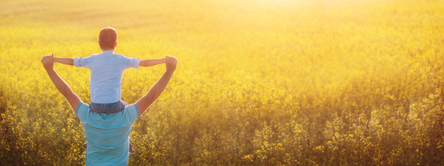 Father holding his son on the shouldders and standing on nature summer background. Back view.
