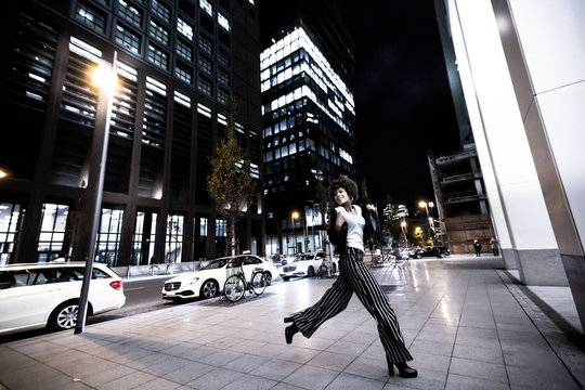 Stylish african american woman walking in night city. Houses illuminated, machines and bicycles on the background. Nightlife in Frankfurt. Evening city walks. Lifestyle concepts