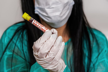 Coronavirus COVID-19 world outbreak concept with vaccine tag on vacutainer. Female chemist with protective uniform, mask and white gloves displays blood tube with Wuhan virus vaccine trial sample.