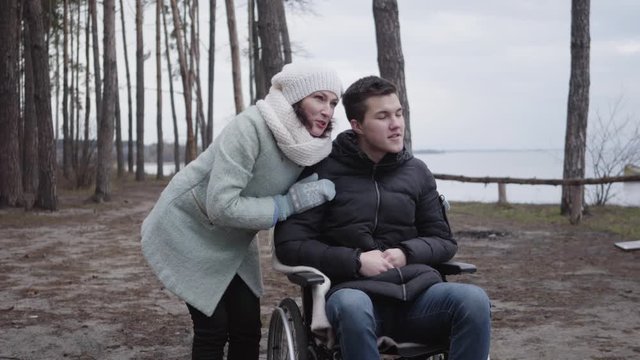 Positive Caucasian man in wheelchair and adult woman looking away in forest on river bank. Portrait of invalid tender or mother enjoying autumn day with crippled son or patient outdoors.