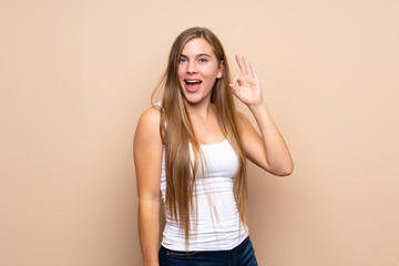 Teenager blonde girl over isolated background surprised and showing ok sign