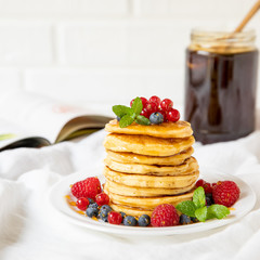 Stack of pancakes with fresh red currant, blueberries, raspberries and honey on a light background for the tasty breakfast.