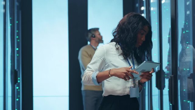 Afro-american young IT specialist woman using digital pad opening rack door of server cabinet analyzing statistics information cooperating in data center.
