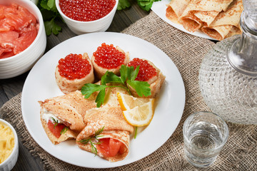 Beautifully rolled pancakes(blini) with fresh sour cream, red caviar and red fish on a white plate. Around a plates with fish, caviar, and fresh butter. Nearby is a decanter, and a stack of vodka.