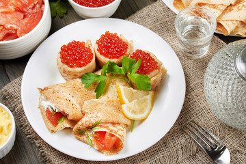 Thin pancakes(blini) with red caviar, red fish and a sprig of parsley and lemon lie on a white plate. Holiday. Maslenitsa.  Rustic style, close up view