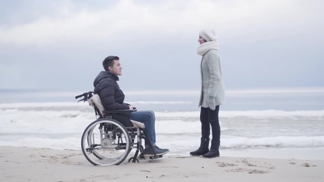Cheerful Caucasian woman talking with boy in wheelchair and pointing away. Adult mother and young disabled son spending autumn day on sea shore outdoors. Leisure, lifestyle, rehabilitation.