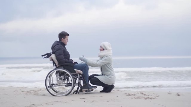 Positive adult Caucasian woman coming to boy in wheelchair and sitting on hunkers. Positive mother spending autumn day with disabled son on sea shore or river bank. Leisure, care, lifestyle.