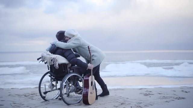 Smiling young mother clapping hands and hugging disabled son in wheelchair. Brunette man playing guitar for parent on sea shore. Support, rehabilitation, care, lifestyle, hobby.