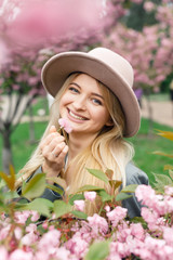 young beautiful blonde girl in a white dress, a gray cloak and a pink hat stands in a park near sakura trees