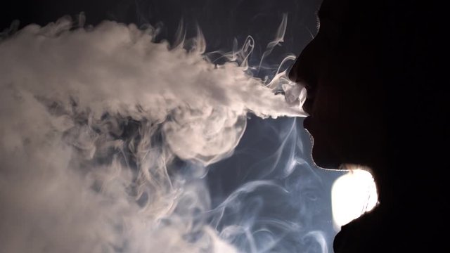 Slow motion isolated silhouetted closeup profile view of a man smoking and blowing out a lot of smoke in dark background