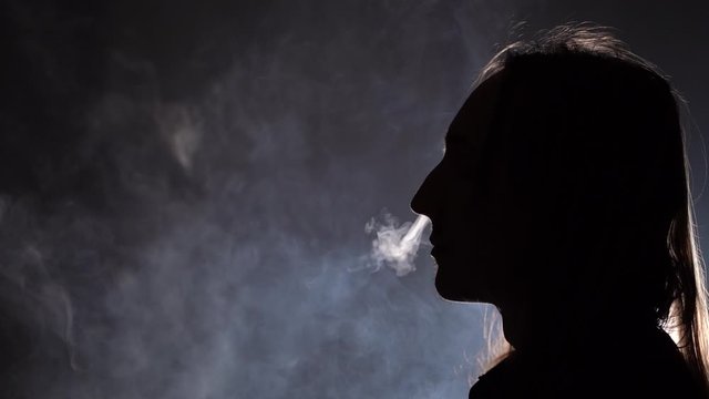 Isolated silhouetted closeup side view of a man with long hair smoking in a dark room blowing smoke through nose