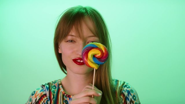 Young woman with adorable smile hypnotizing with the colorful lollipop. Slow motion shot isolated on the green background