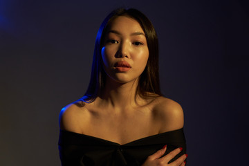 Fototapeta premium Portrait of a young beautiful Asian girl looking directly at the camera. The woman's hair and shoulders in colorful bright UV blue lights. On black background. 
