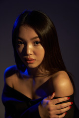 Portrait of a young beautiful Asian girl looking directly at the camera. The woman's hair and shoulders in colorful bright UV blue lights. On black background.  