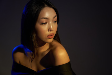 Portrait of a young beautiful Asian girl looking away. The woman's hair and shoulders in colorful bright UV blue lights. On black background.   